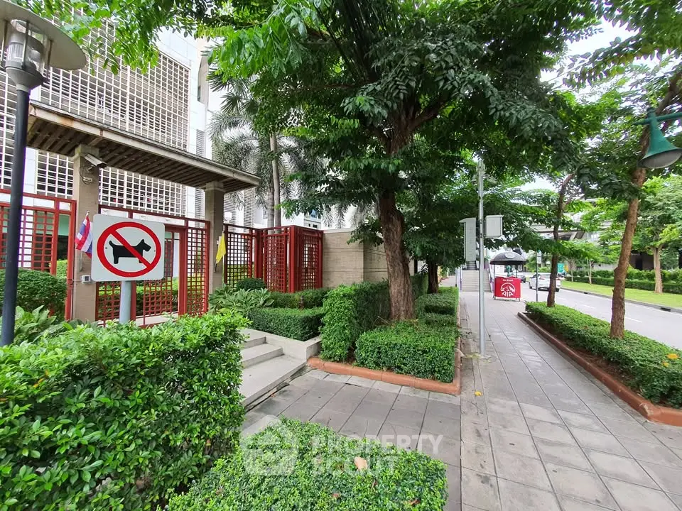 Lush green entrance to modern building with red accents and paved walkway