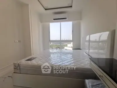 Bright bedroom with large window and city view, featuring a new mattress and air conditioning unit.