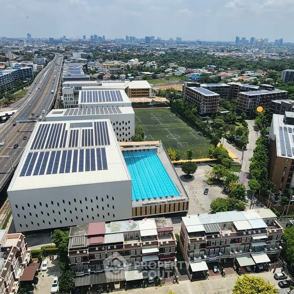 Aerial view of modern building complex with solar panels, pool, and sports field in urban setting.