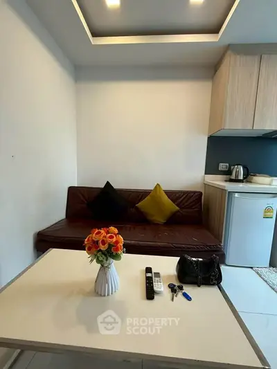 Cozy living room with modern decor, featuring a compact sofa, stylish table, and kitchenette with fridge and kettle.