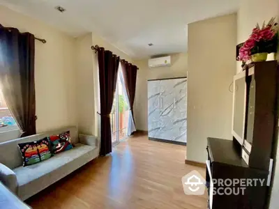 Sunlit spacious living room with elegant hardwood flooring, cozy sofa, and large windows draped in dark curtains, leading to a balcony.