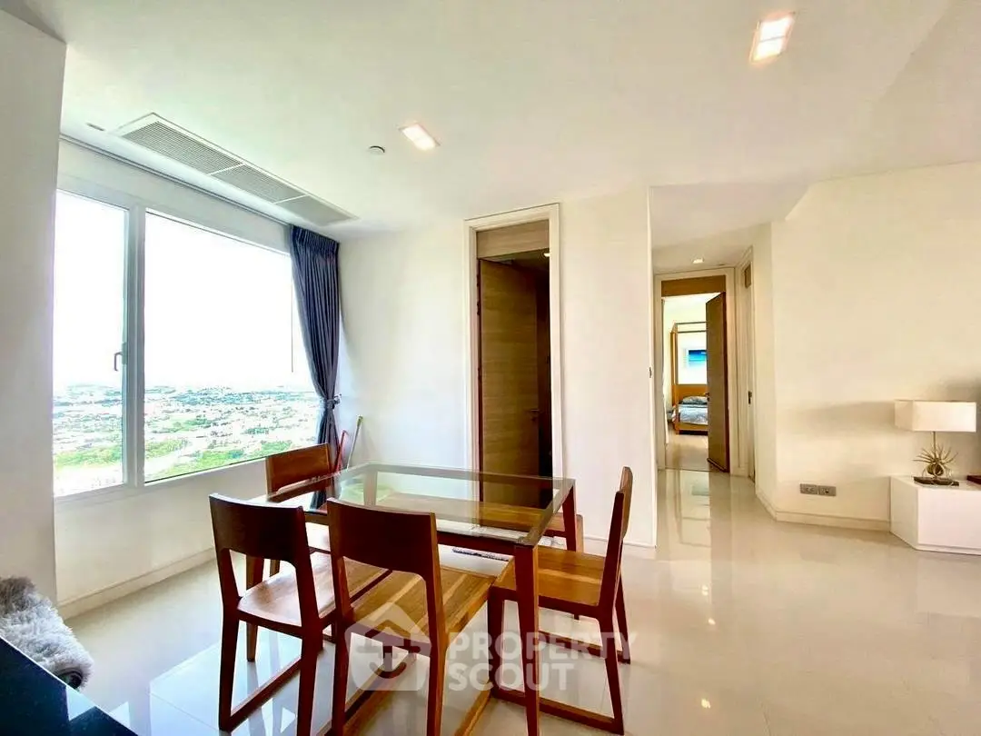 Bright dining area with glass table and wooden chairs, offering stunning views through large window.