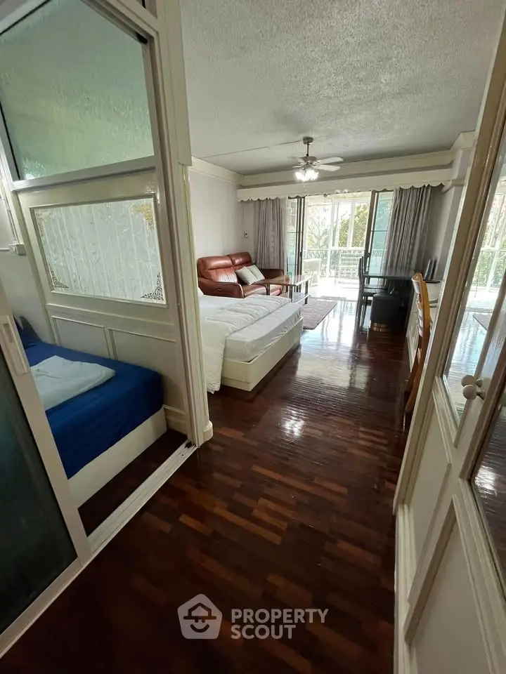 Spacious living room with wooden flooring and large windows opening to a balcony, featuring cozy seating and natural light.