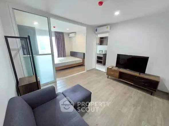 Modern living room with sleek furniture and open view into bedroom, featuring contemporary design and ample natural light.