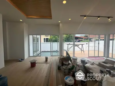 Spacious living room under renovation with large windows and balcony access, featuring modern lighting and wooden flooring.