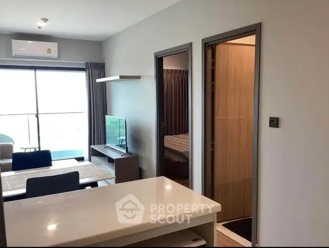 Modern apartment interior with an open-plan living space featuring a sleek white table, cozy sofa, and a flat-screen TV, bathed in natural light from large windows.