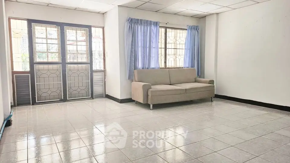 Spacious living room with tiled floor and natural light, featuring a comfortable sofa and large windows.