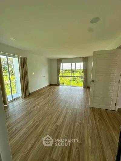 Spacious empty room with large windows and wooden flooring, offering a serene view of greenery.