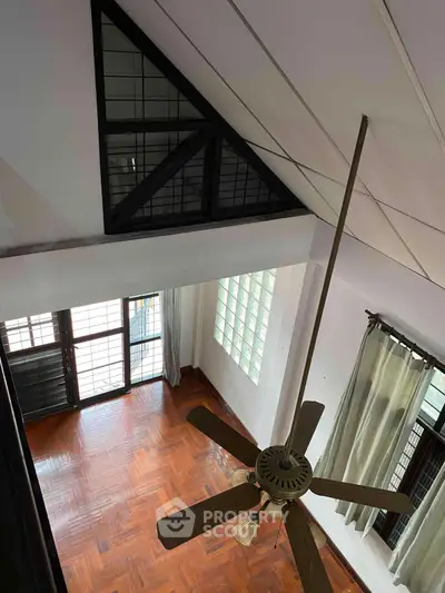 Spacious living room with high ceiling and wooden flooring, featuring a ceiling fan and large windows for natural light.