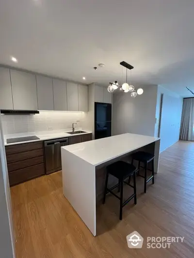 Sleek modern kitchen with white countertops, stylish lighting, and integrated appliances, opening into a spacious living area with hardwood floors.