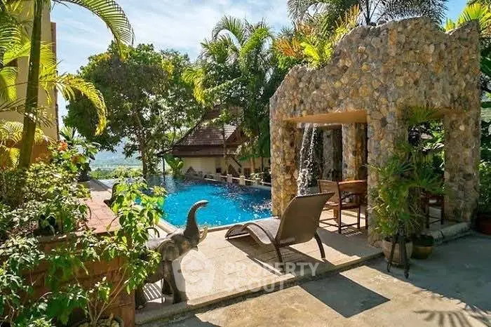 Luxurious tropical poolside retreat with lush greenery and stone waterfall feature.