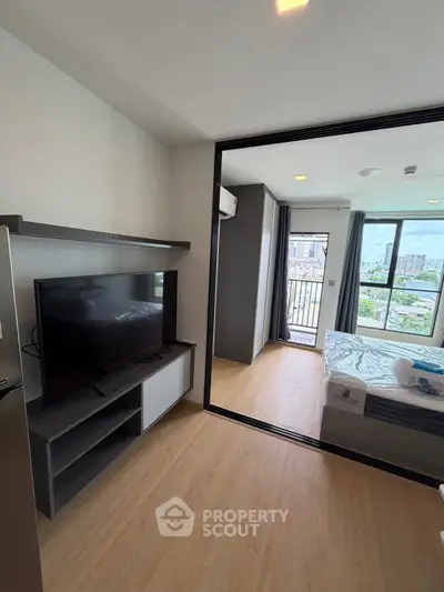 Modern apartment interior with open living room and bedroom, featuring a large TV and city view balcony.