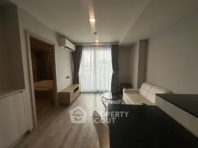 Modern living room with sleek furniture and natural light from large window, adjacent to a cozy bedroom.