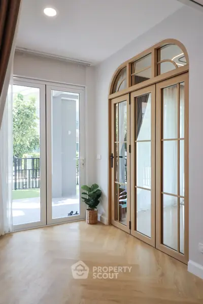 Bright living room with elegant wooden doors and large windows opening to a balcony with lush greenery.
