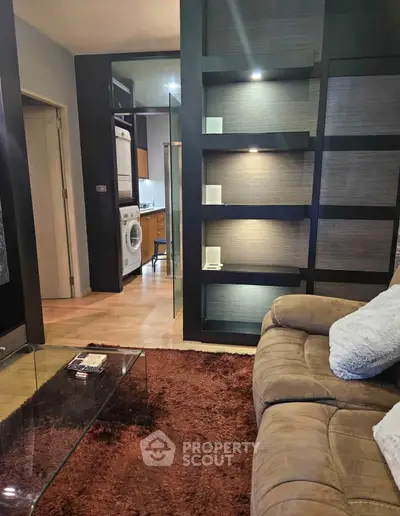 Modern living room with cozy sofa, glass coffee table, and view into kitchen with washing machine.