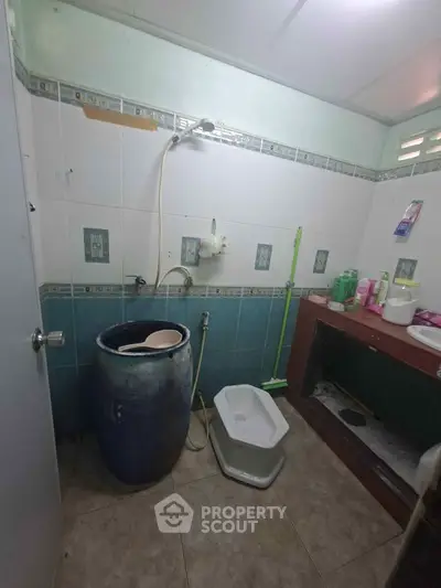 Traditional bathroom with squat toilet and water storage barrel, featuring tiled walls and floor.