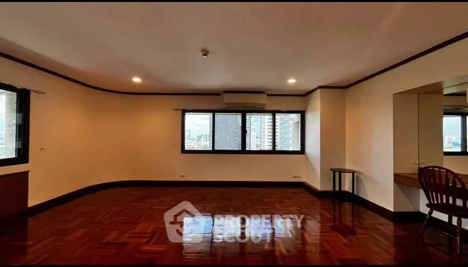Spacious empty room with polished wooden floors and city view windows.