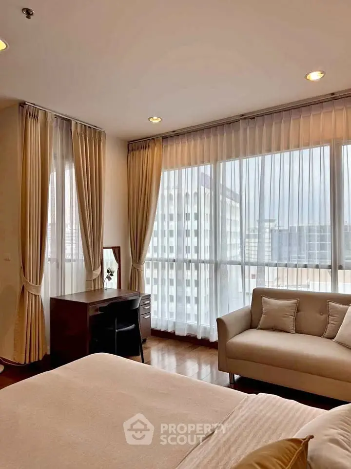 Spacious bedroom with large windows offering city views, elegant curtains, and cozy seating area.