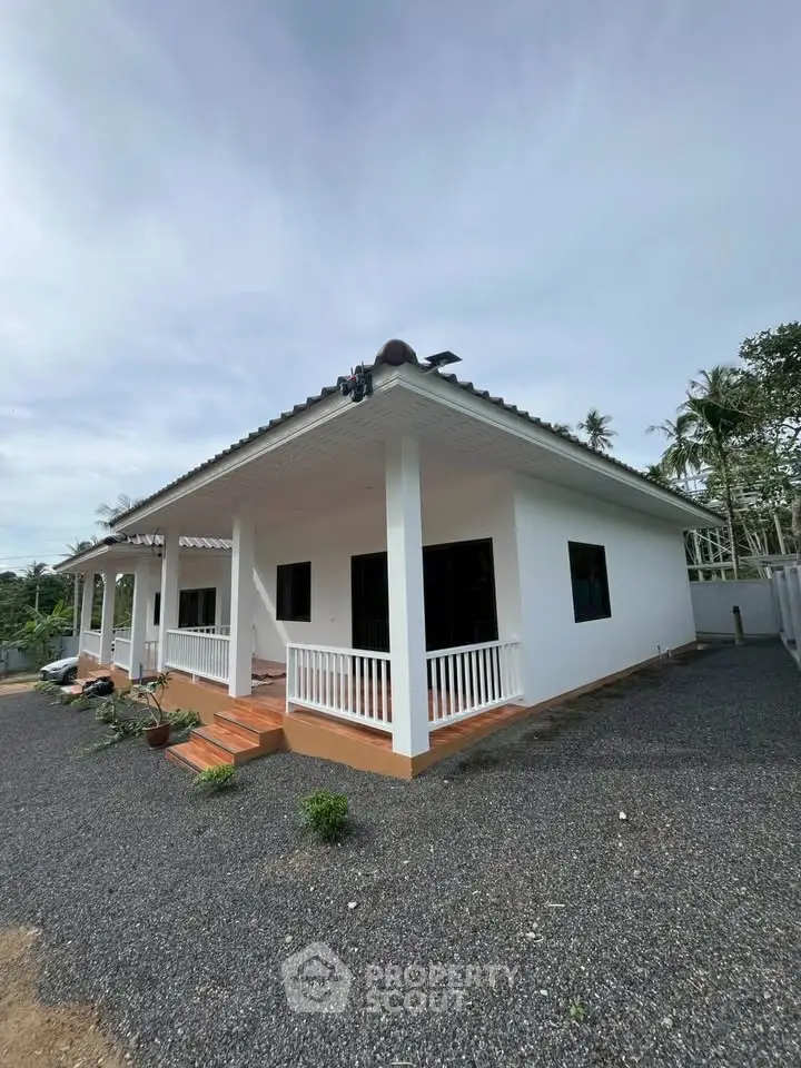 Charming single-story white house with a spacious porch and gravel driveway, surrounded by lush greenery.