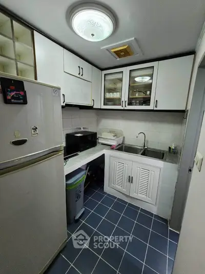 Compact kitchen with white cabinets, double sink, and tiled floor, featuring a fridge and microwave.