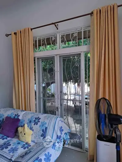 Cozy living room with floral sofa, large window, and elegant curtains, offering a glimpse of the garden outside.