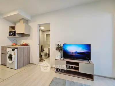 Modern open-plan living room with compact kitchen and washing machine, featuring a flat-screen TV and stylish decor.