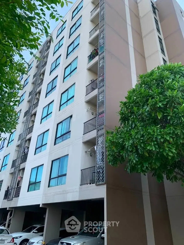 Modern multi-story apartment building with balconies and parking area, surrounded by lush greenery.