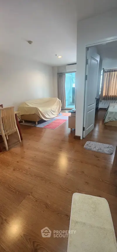 Spacious living area with wooden flooring and natural light, featuring a cozy seating arrangement and bedroom access.