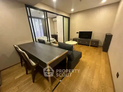 Modern living room with wooden flooring, sleek furniture, and a view into the bedroom through glass doors.