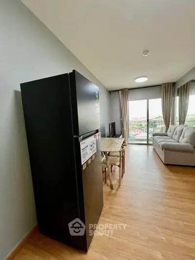 Modern living room with sleek black fridge and cozy seating area, featuring large windows and wooden flooring.