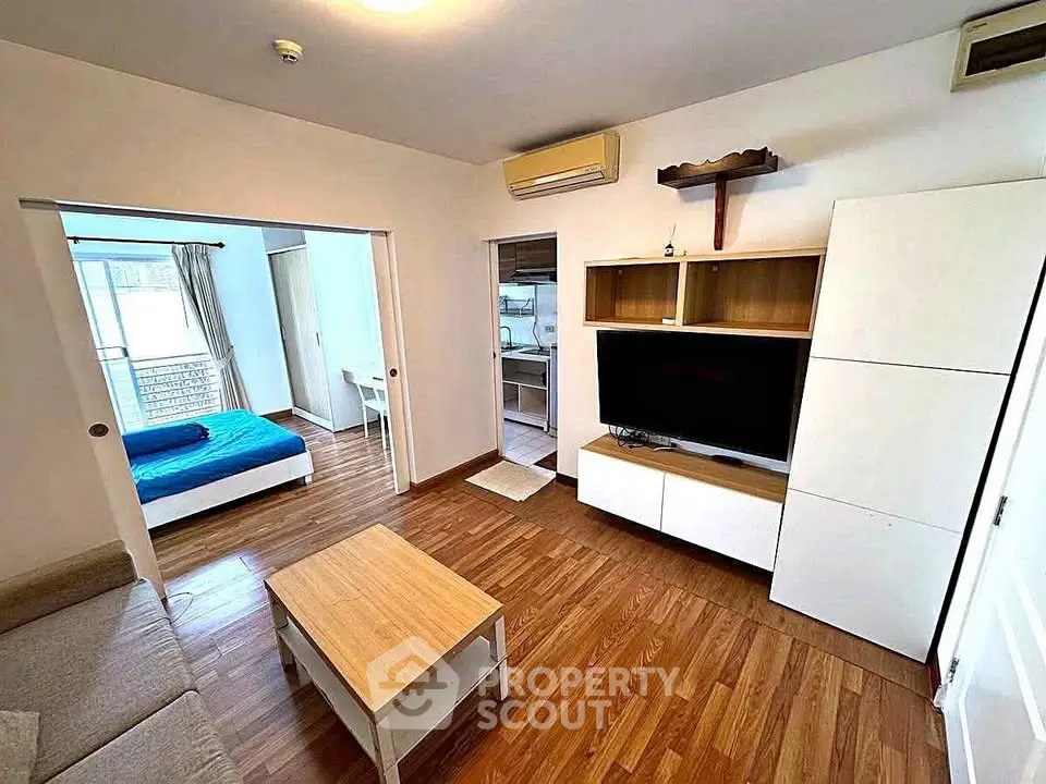 Modern living room with wooden flooring, TV unit, and view into a cozy bedroom.