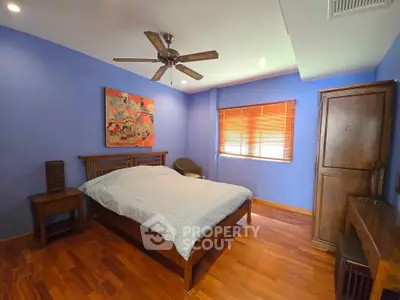 Spacious bedroom with wooden flooring and ceiling fan, featuring vibrant blue walls and cozy furnishings.