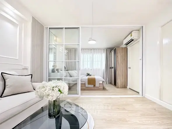 Modern living room with glass partition leading to a cozy bedroom, featuring elegant decor and natural light.