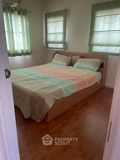 Cozy bedroom with wooden flooring and pastel bedding, featuring natural light through green curtains.
