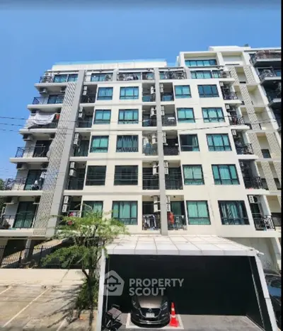 Modern multi-story apartment building with balconies and parking area under clear blue sky.