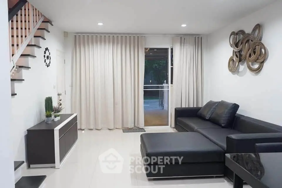 Modern living room with sleek black furniture and elegant decor, featuring a staircase and large window with curtains.