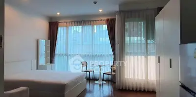 Modern bedroom with large windows and city view, featuring a cozy sitting area and sleek furnishings.