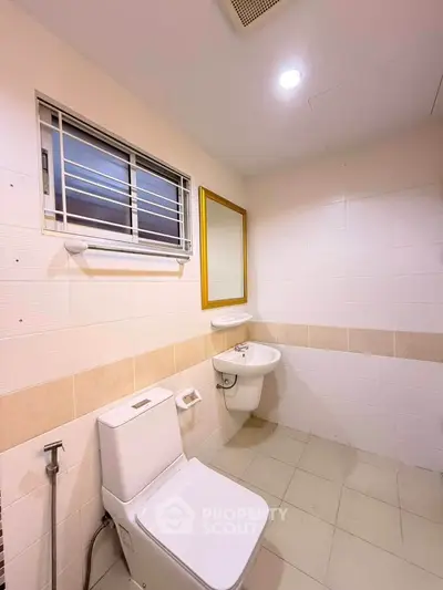 Modern bathroom with white tiles, toilet, and sink, featuring a window and mirror for natural light.