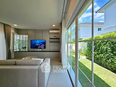 Modern living room with large windows overlooking a lush garden, featuring sleek furniture and a wall-mounted TV.