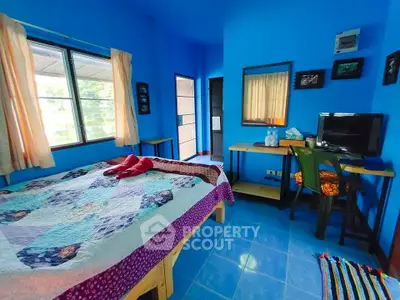 Cozy bedroom with vibrant blue walls and natural light, featuring a desk and TV.