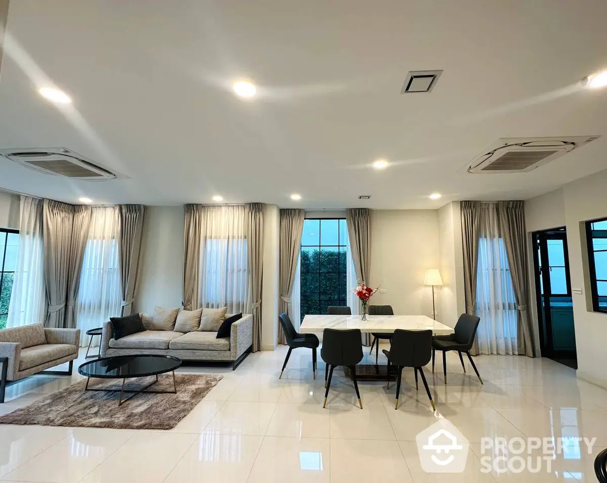 Spacious and elegantly designed living room with modern furniture, sleek lighting, and large windows draped in luxurious curtains, leading to a cozy dining area.