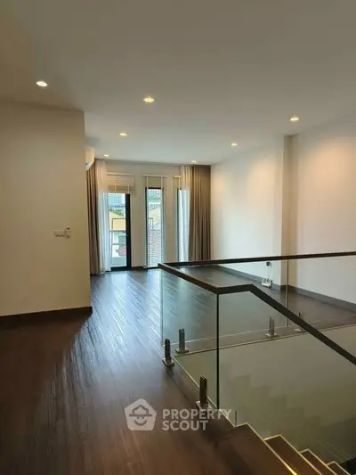 Spacious modern living room with wooden flooring and glass railing, featuring large windows for natural light.
