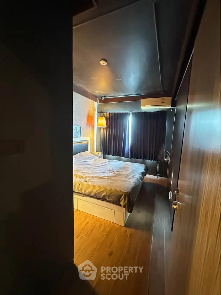 Cozy bedroom with wooden flooring and ambient lighting, featuring a comfortable bed and dark curtains.