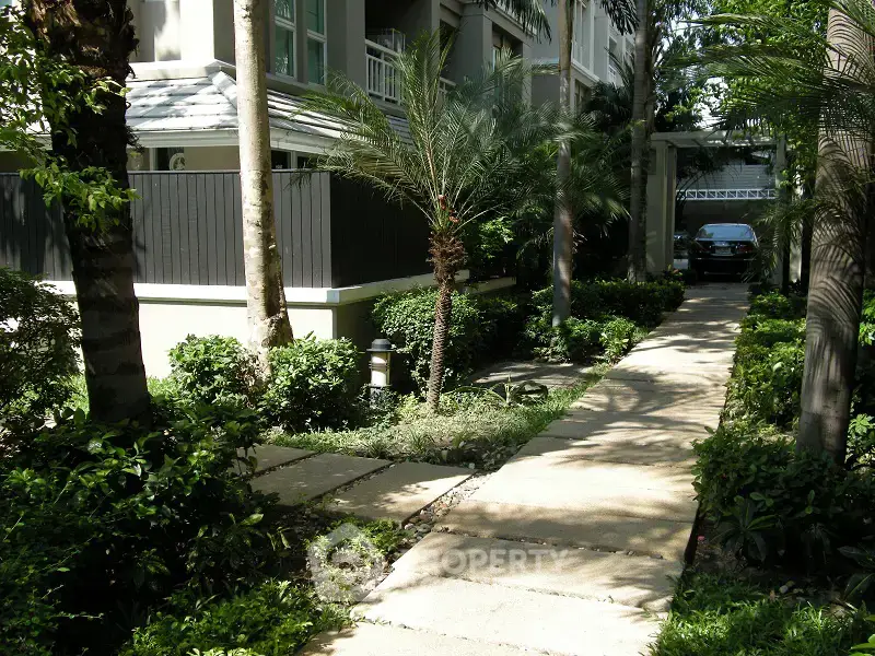 Lush garden pathway leading to a modern residential building entrance, surrounded by greenery and palm trees.