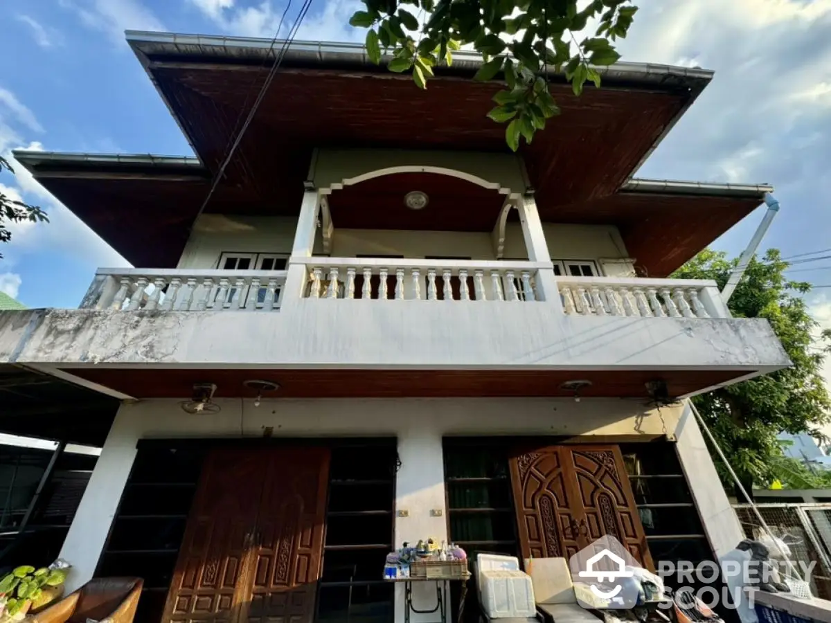 Charming two-story house with a spacious balcony and rustic wooden doors, surrounded by lush greenery.