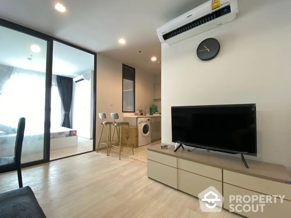 Modern living room with open kitchen and washing machine, featuring sleek furniture and natural light.