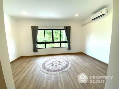 Spacious and bright living room with large windows offering a serene tree-lined view, complemented by a stylish area rug and modern air conditioning unit.