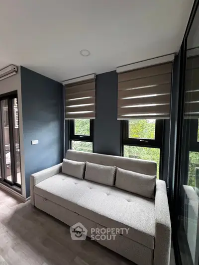 Modern living room with sleek gray sofa and large windows offering lush green views.