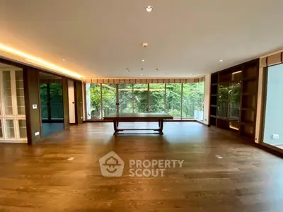 Spacious living room with large windows offering lush green views, featuring elegant wooden flooring and built-in shelves.