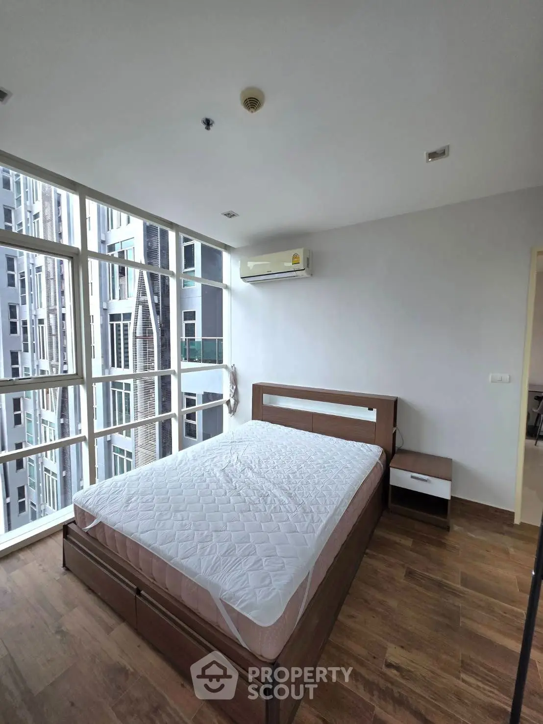 Modern bedroom with large windows and city view, featuring wooden flooring and minimalist design.